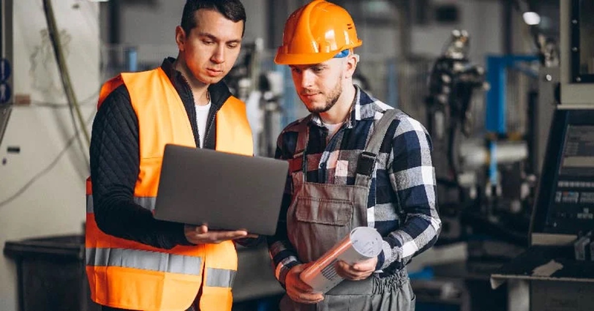 Two industrial workers in hi-vis vest and hard hat review a laptop on the factory floor
